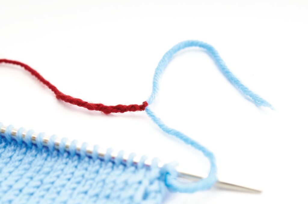 Hellblaues Strickprojekt mit silberner Nadel, Kontrastgarn in Rot. Handarbeit, diy, modisch.